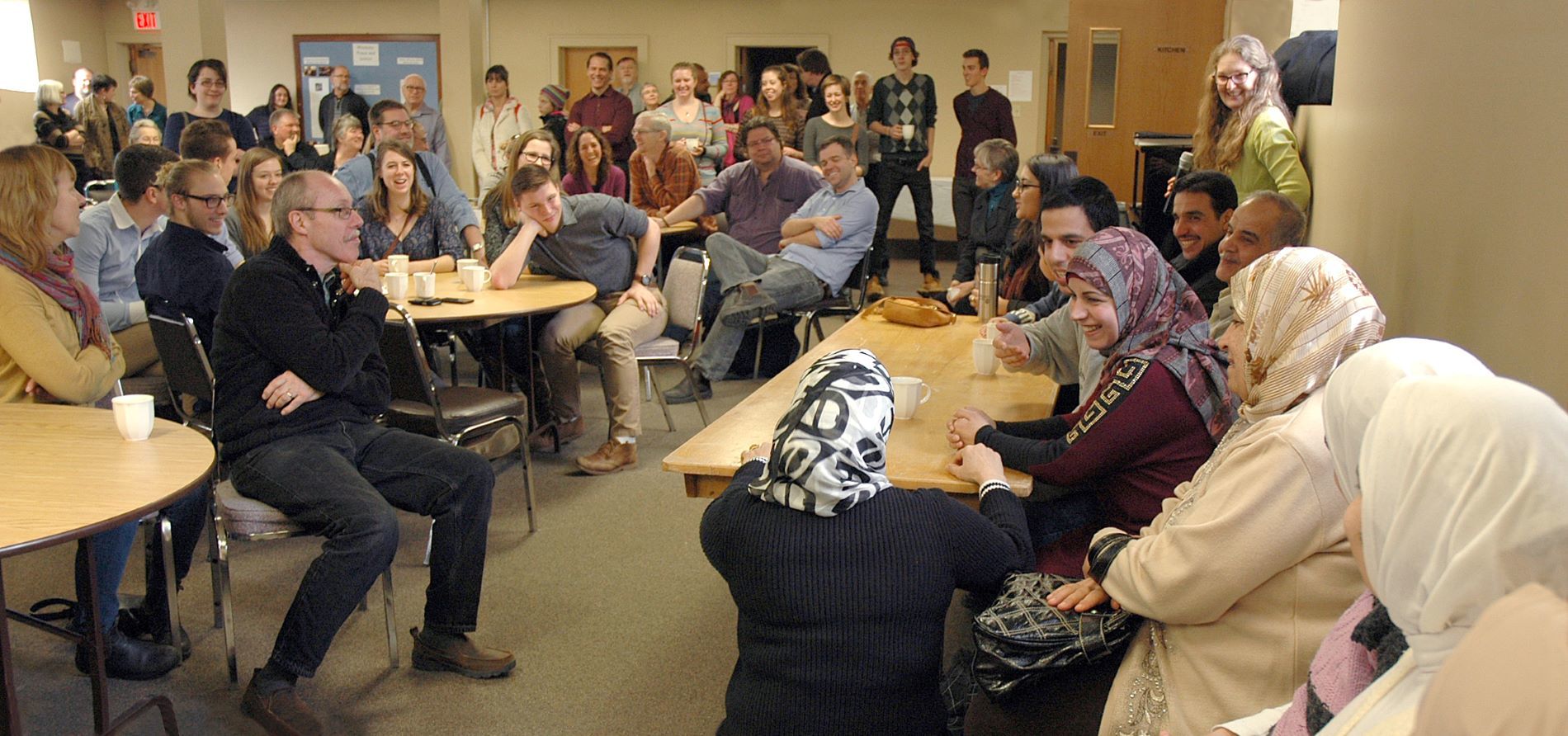 church members and Syrian friends gather in basement of church standing and around tables